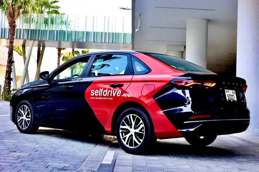 Black and red car with selfdrive.app logo parked on gray brick road, palm trees and building in background, showcasing car subscription service with white text on side panel, sleek design and silver rims visible.