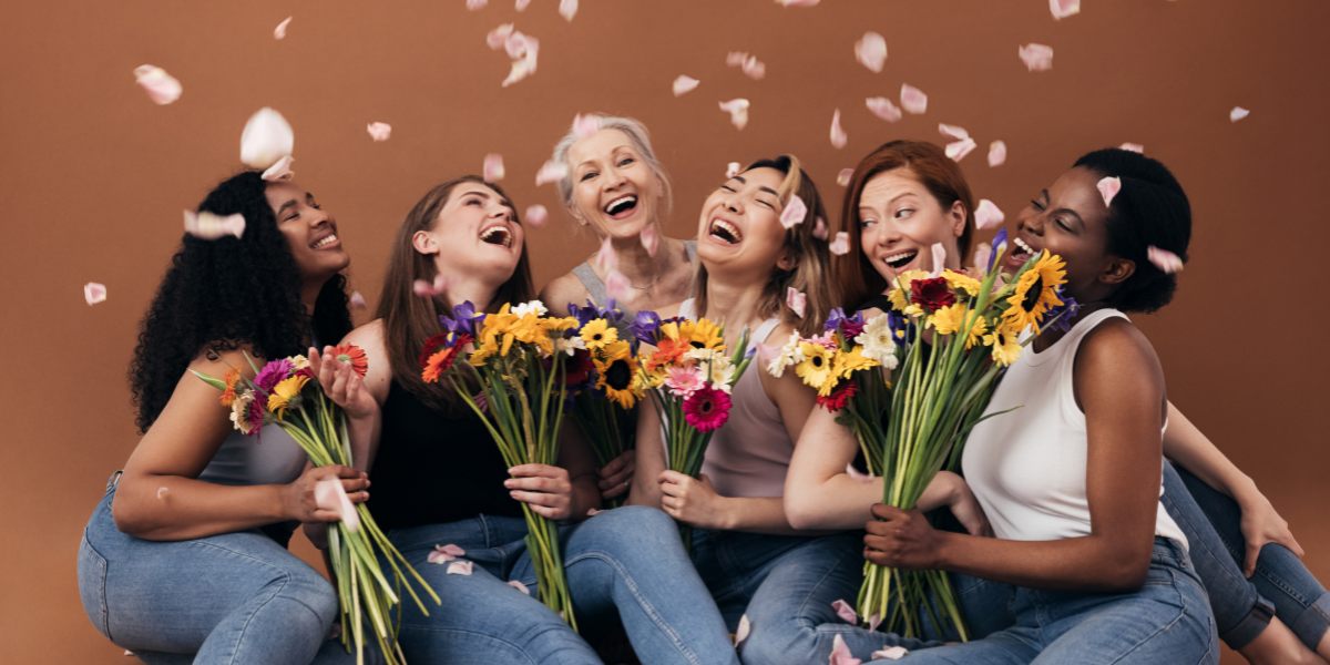 A group of six women of diverse backgrounds joyfully hold colorful bouquets against a brown background, with petals falling around them, conveying happiness and unity.